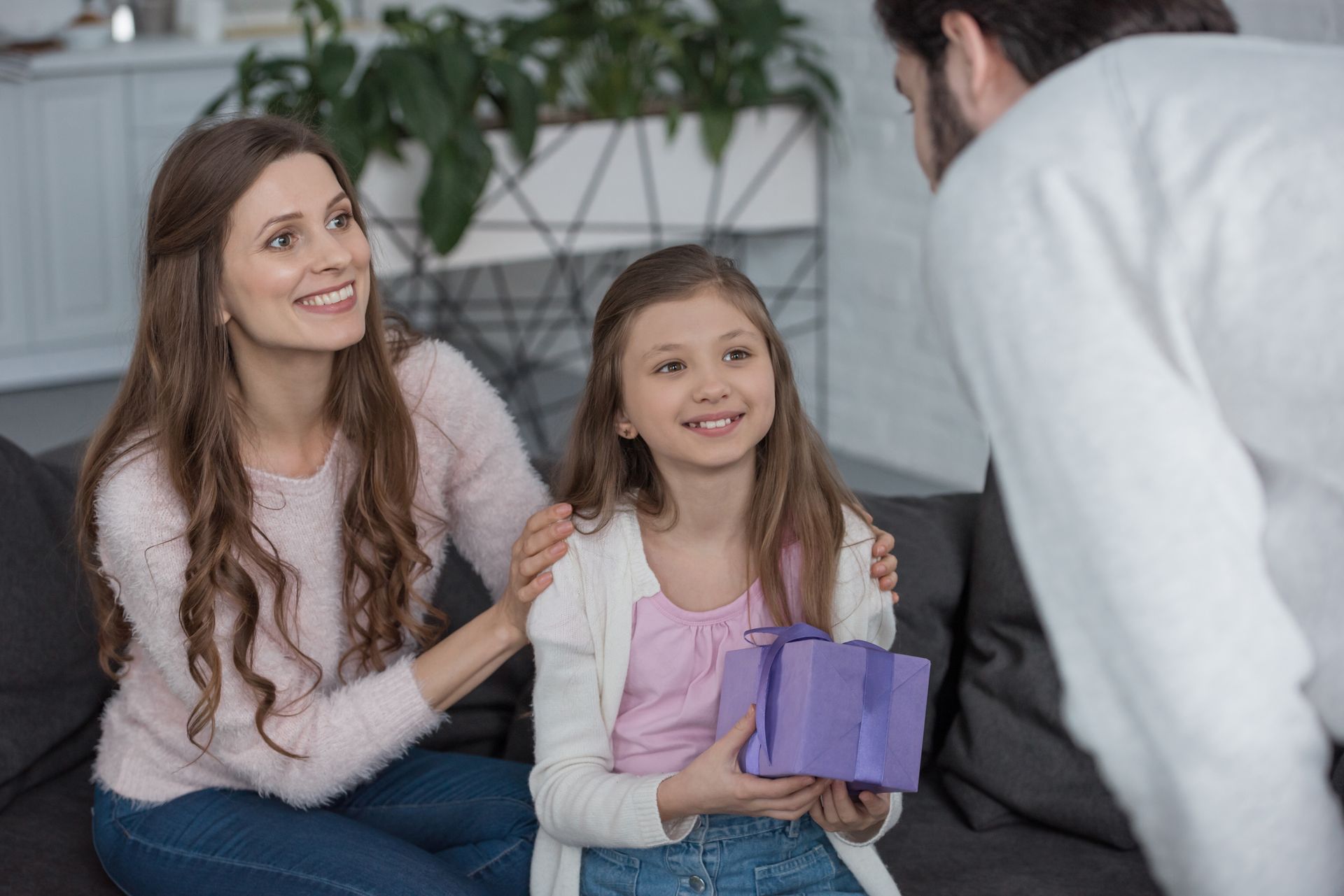 Woman and smiling child holding a purple gift box while a man leans in to talk to them indoors