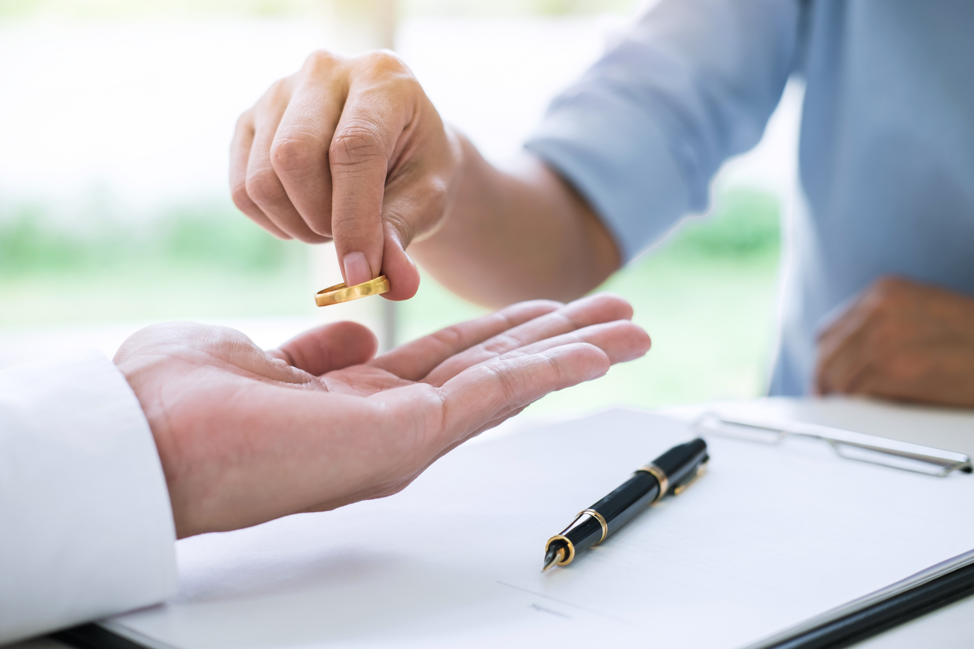 Hand placing a gold ring into an open palm beside a black pen and clipboard on a desk