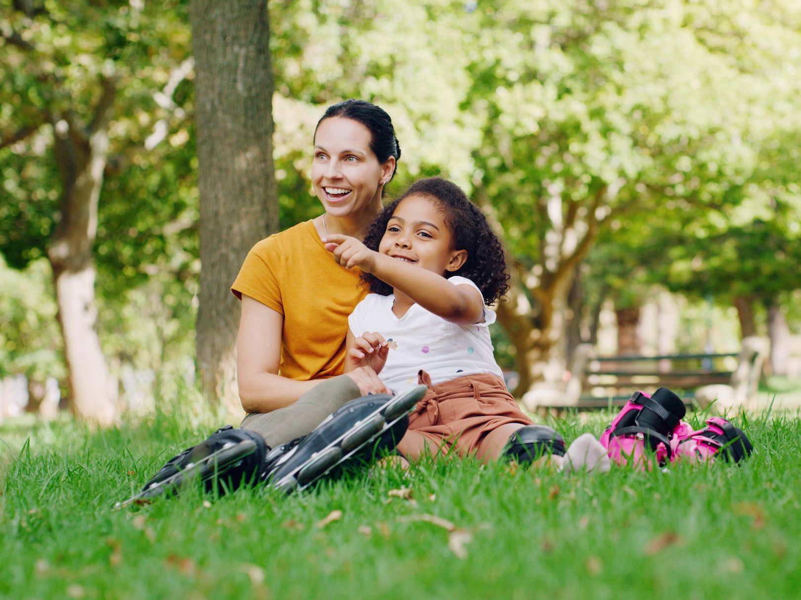 Woman and child sitting on grass in a park, smiling and pointing ahead, with a backpack nearby.