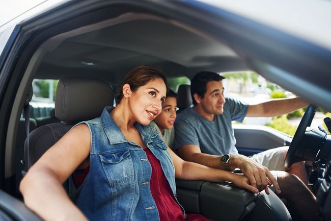 Three people ride in a car, with the driver pointing ahead and a woman smiling in the front passenger seat.