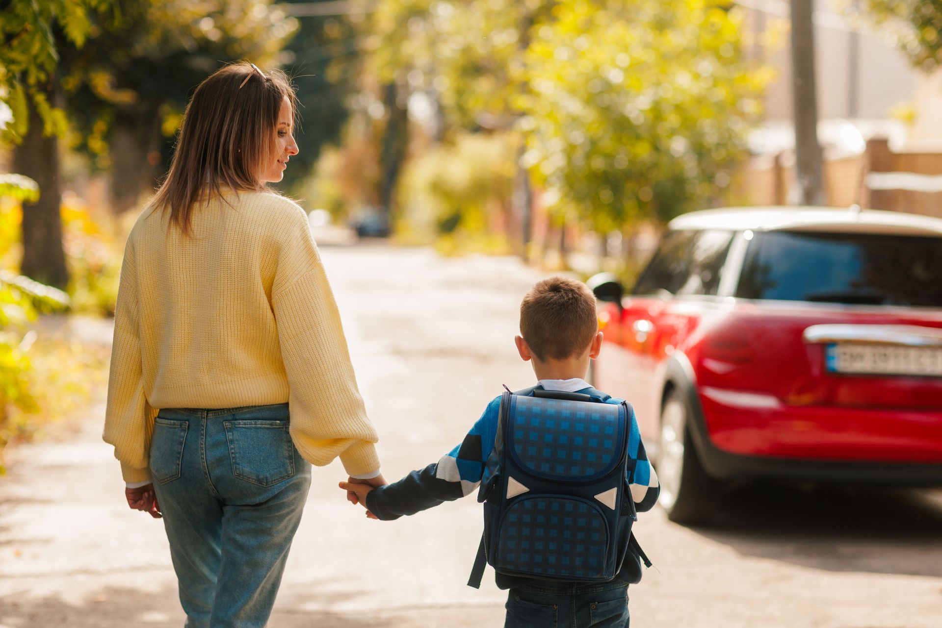 Adult and child holding hands on a sunlit sidewalk beside a red car