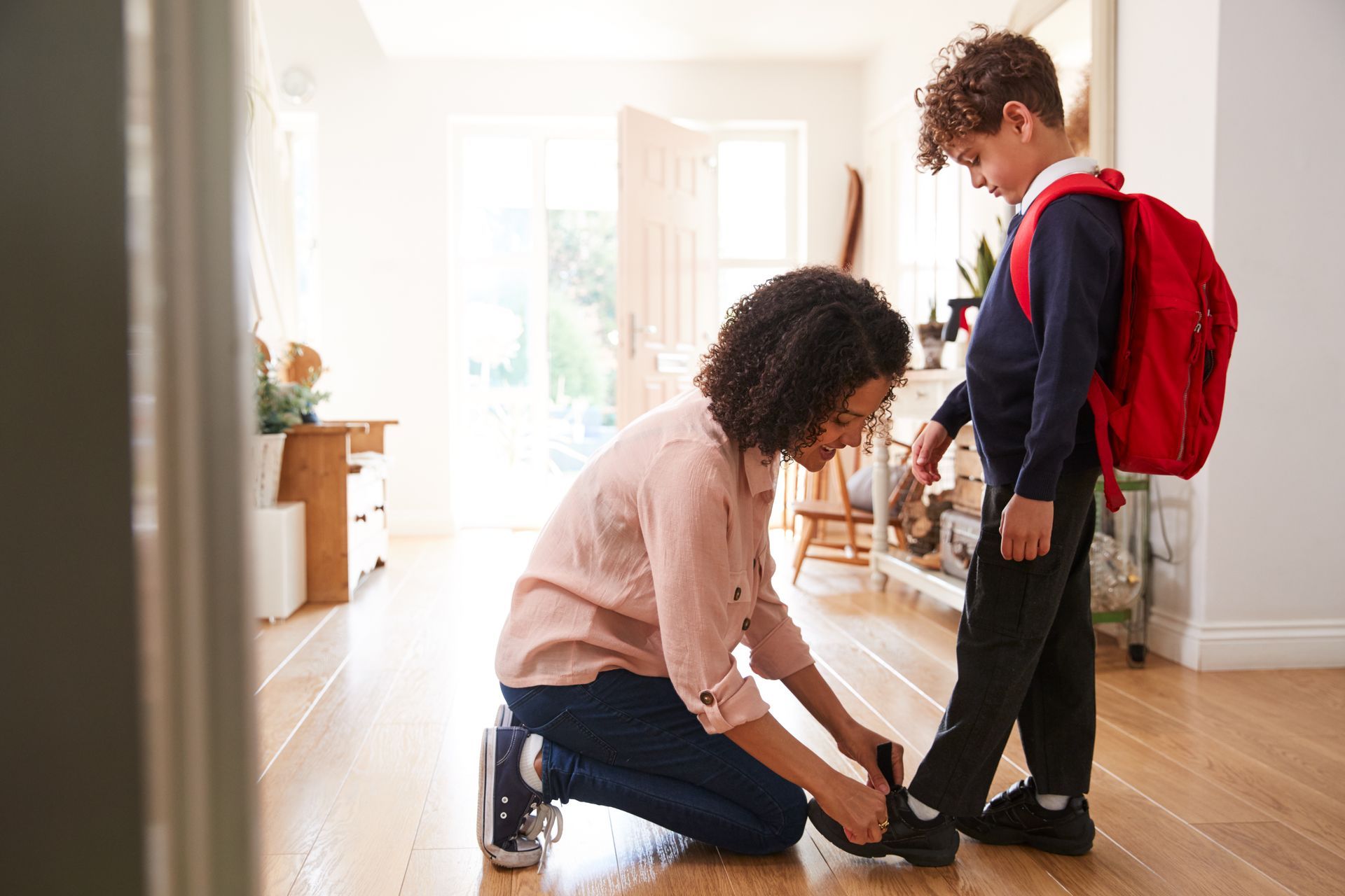 Adult kneeling to tie a child’s shoelace in a bright hallway, with a red backpack nearby.