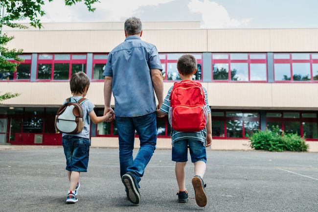 Two children and an adult walking toward a school building with backpacks
