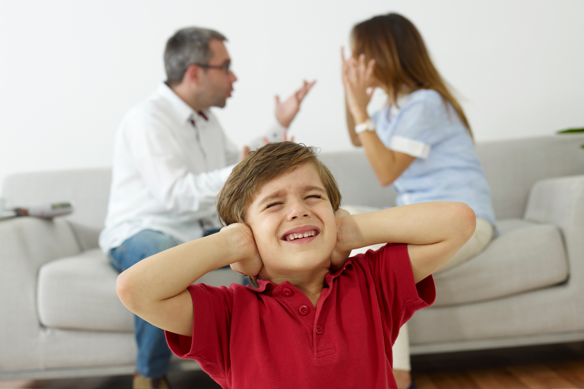 Child covering ears while adults argue on a couch in the background