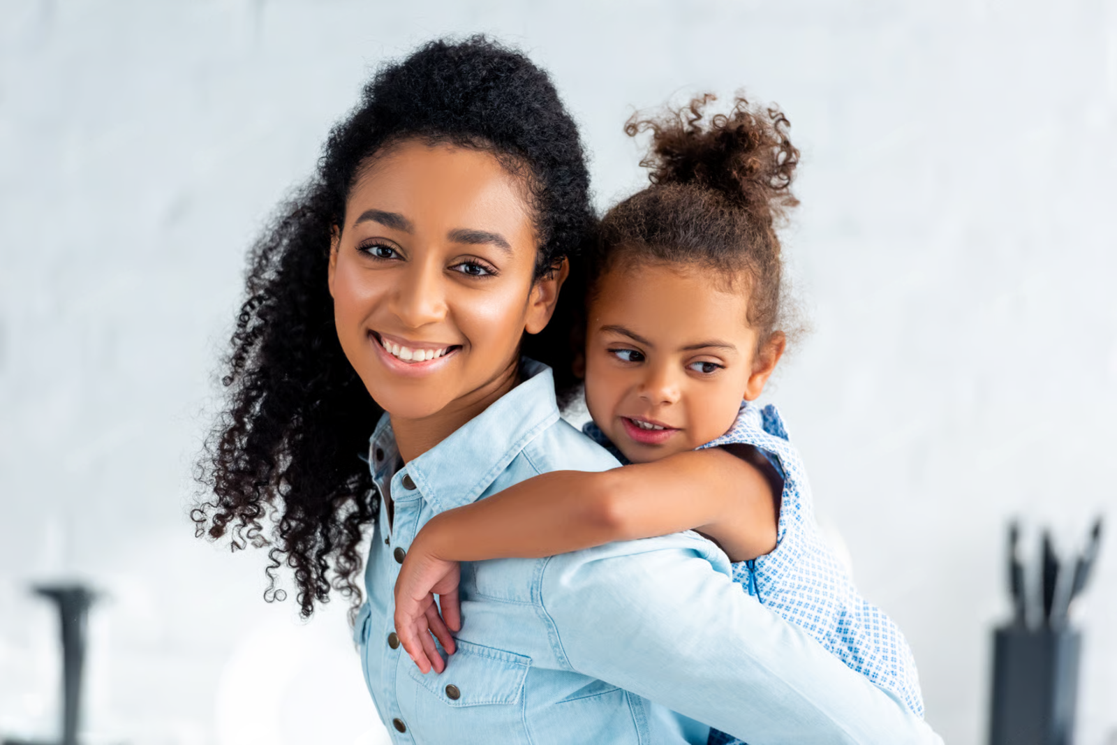 Two smiling people in light blue shirts, one giving the other a piggyback ride indoors.