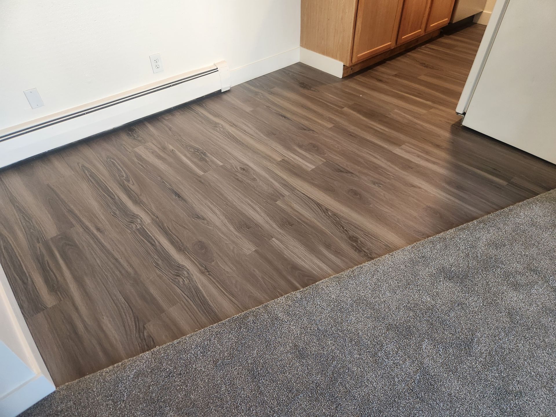 Small room with wood-look flooring, gray carpet edge, white wall heater, and wooden cabinets.