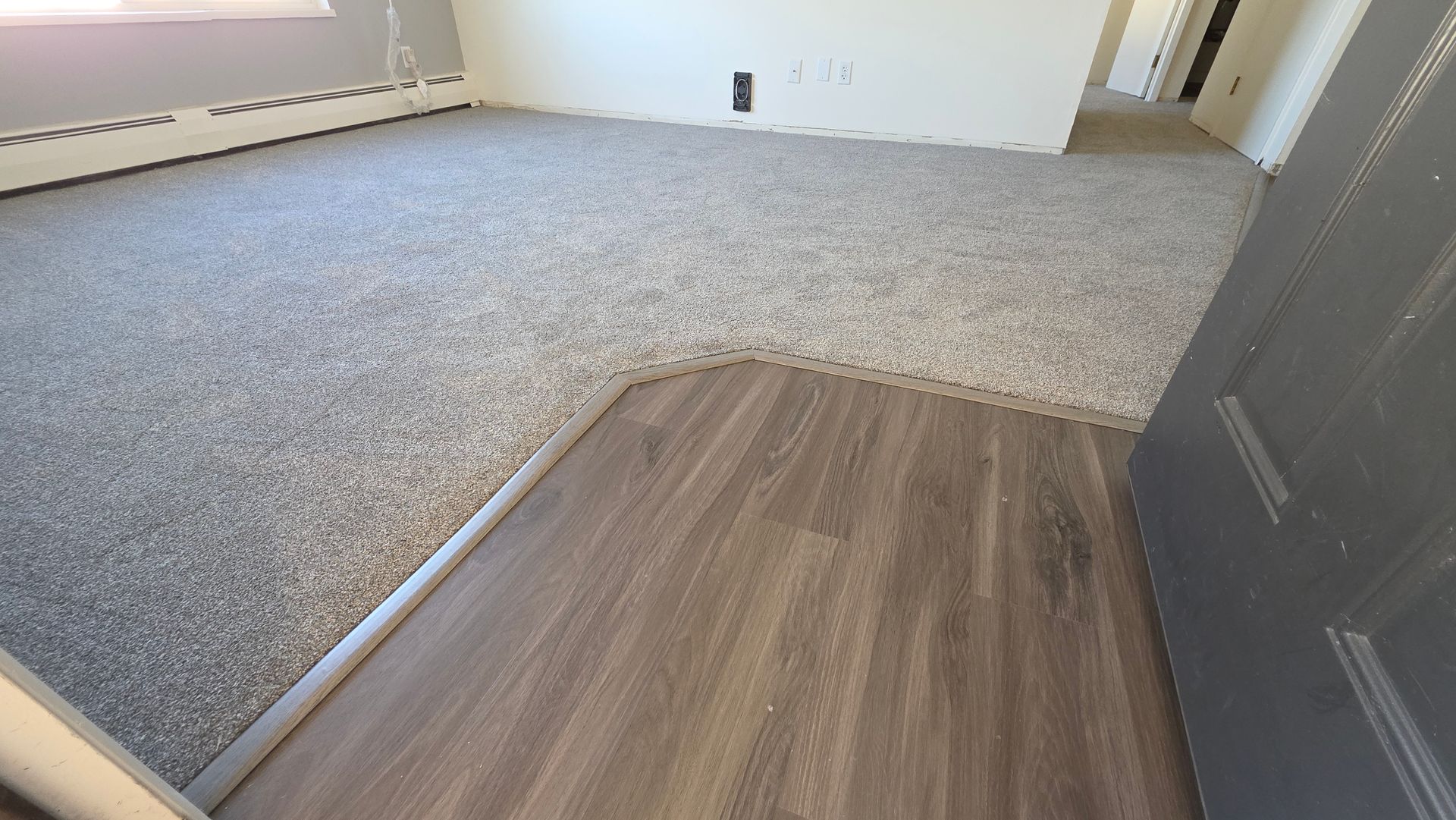 Empty living room with gray carpet, wood flooring entry, and white walls with a doorway.