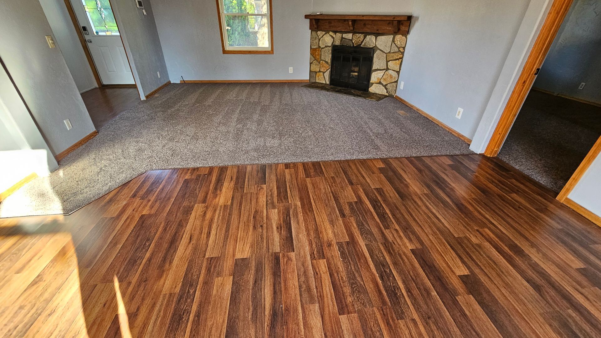 Empty living room with wood floor, gray carpet area, and stone fireplace.