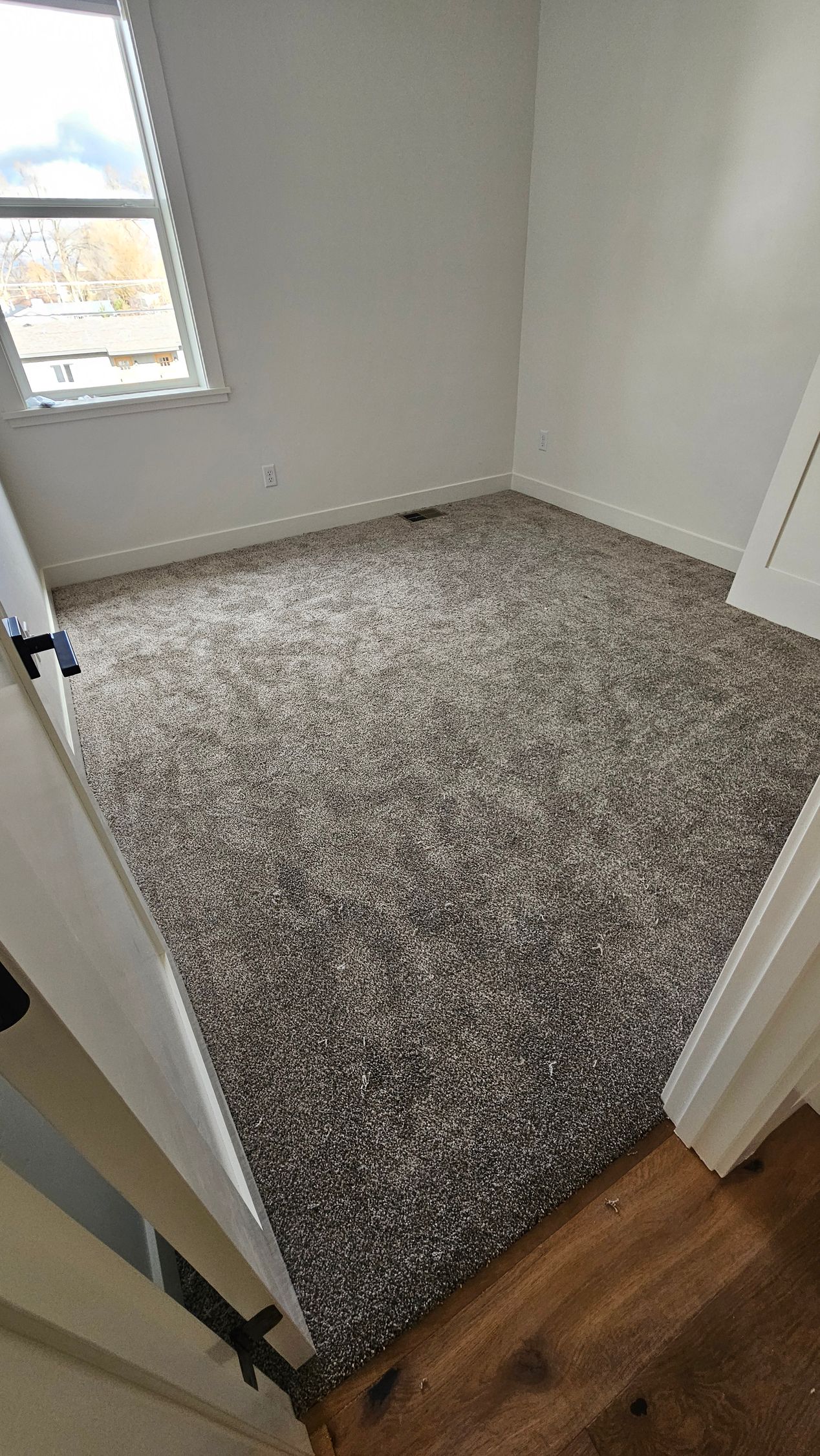 Empty small room with gray carpet, white walls, a window, and wood flooring at the doorway
