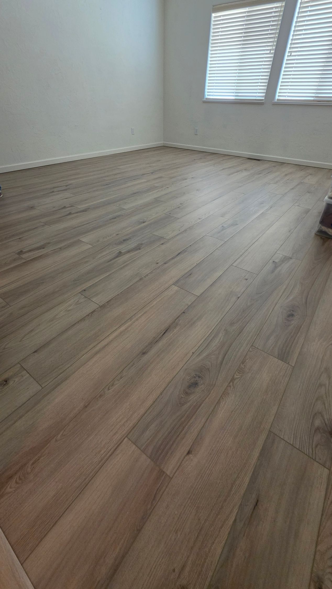 Empty room with light wood flooring and white walls, lit by a window with blinds.