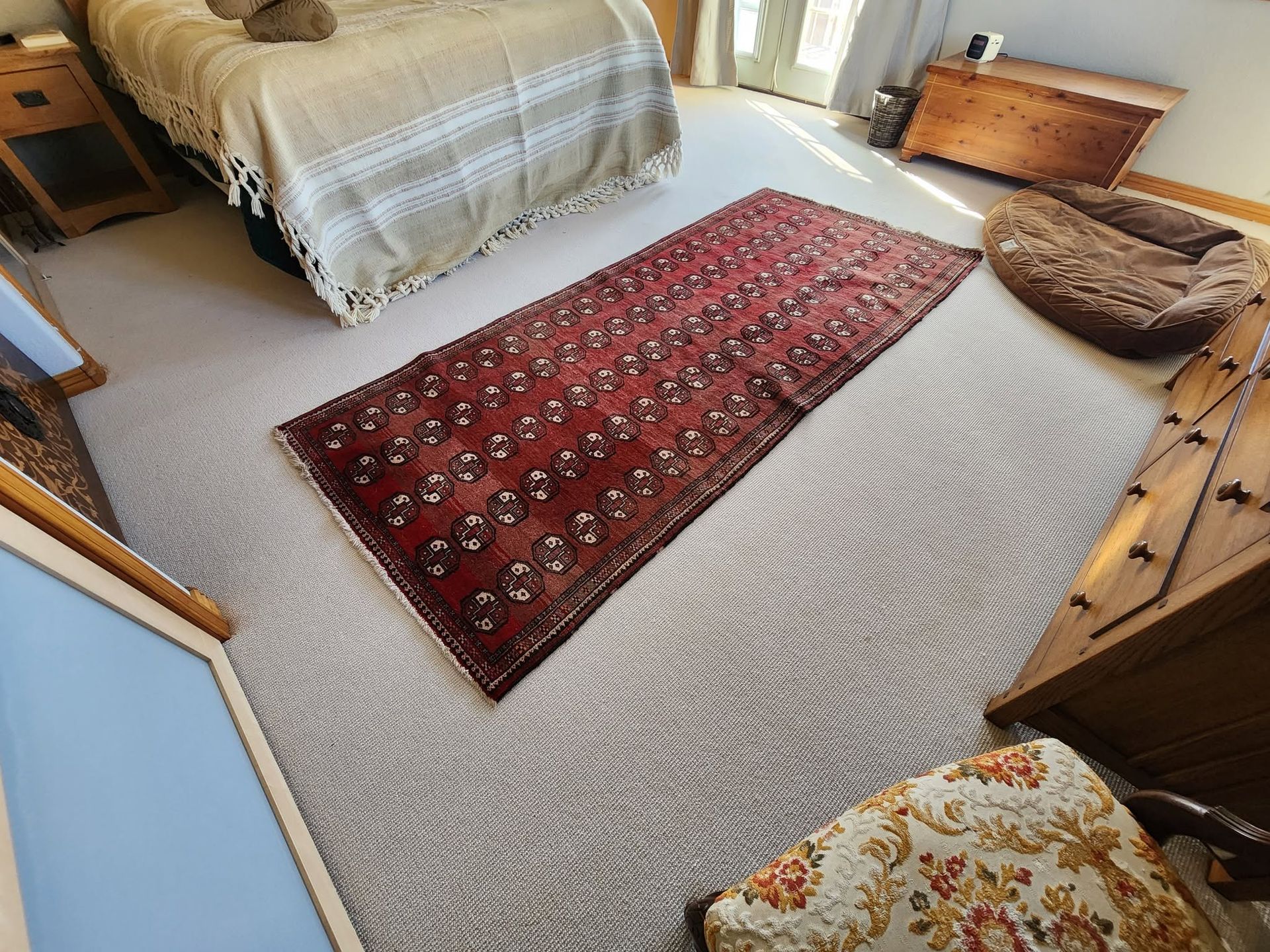 Bedroom with beige carpet, red runner rug, bed, dresser, and wicker pet bed near the window