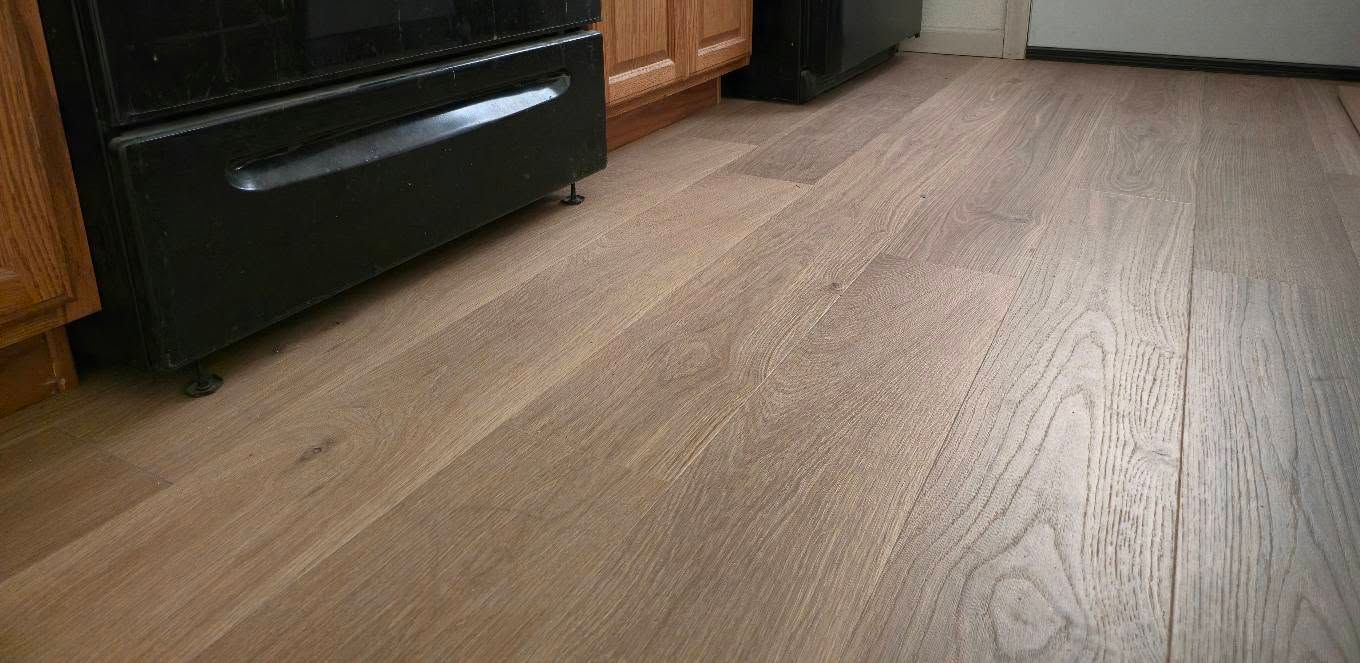 Low-angle view of a kitchen floor with wood-look vinyl and a black oven at left, cabinets in the back.
