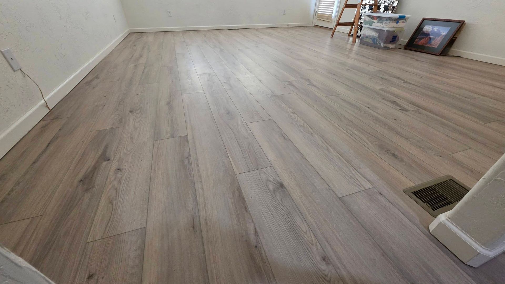 Empty room with light wood flooring, a wall vent, and framed items leaning against the wall.