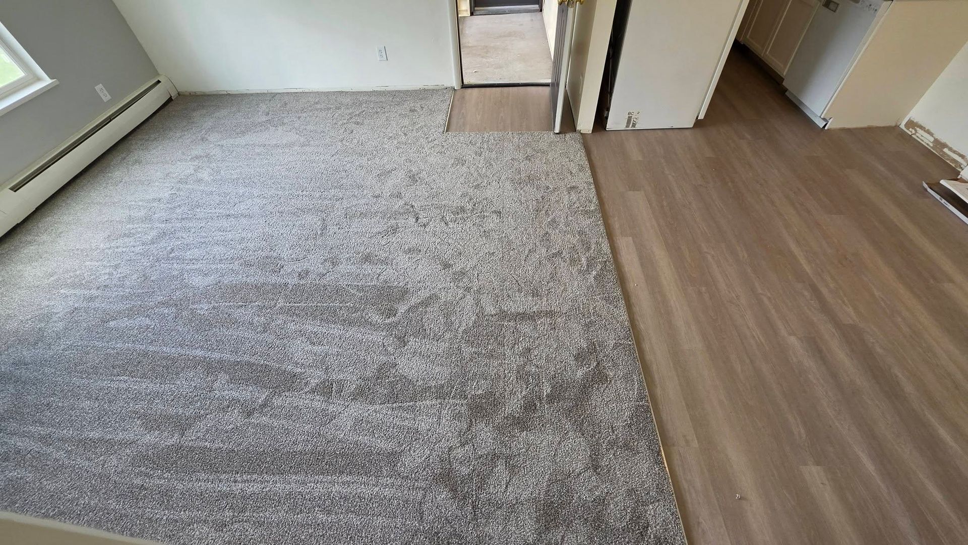Empty room with gray carpet beside light wood flooring and white walls, near a doorway.