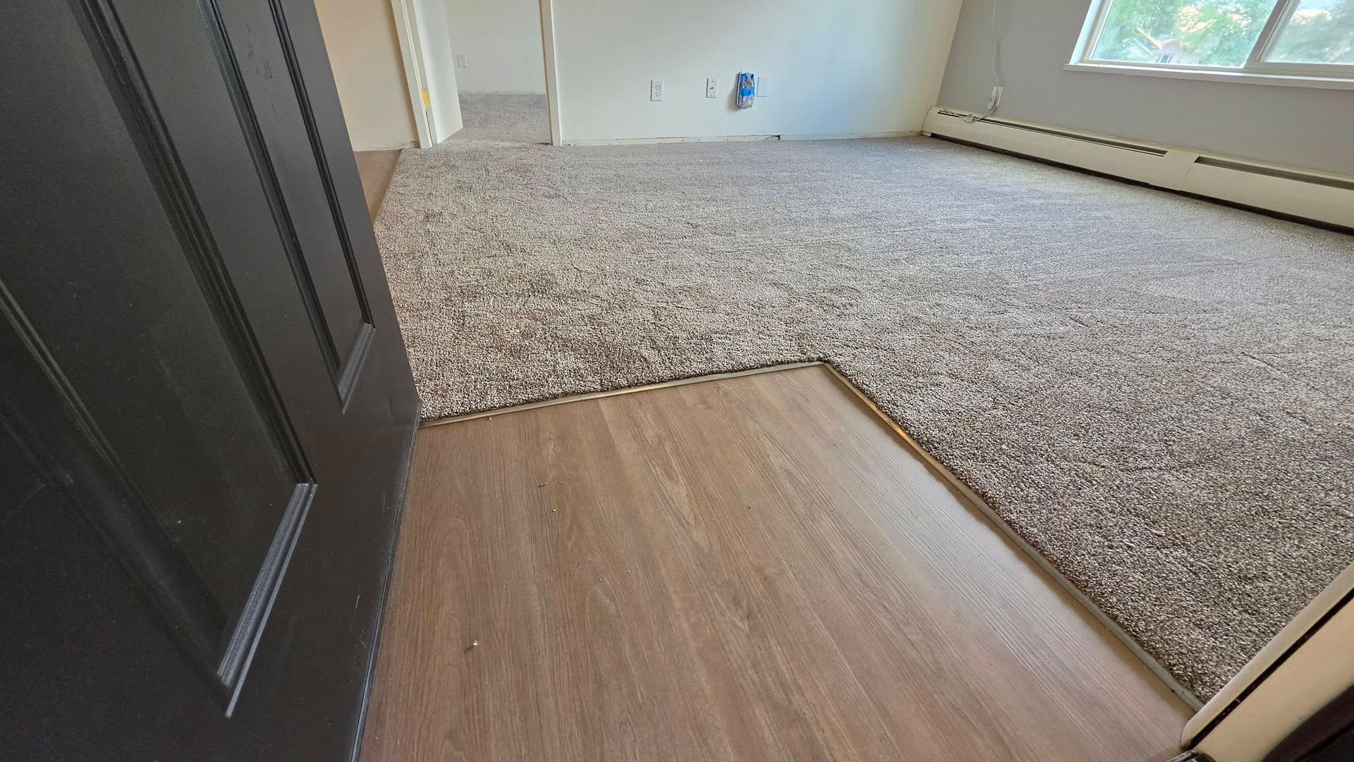 Empty room with carpet and wood flooring transition near a black door, white walls, and a window.