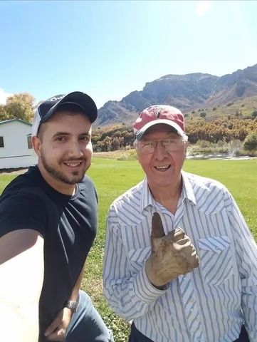 Two smiling men posing outdoors with mountains in the background, one holding up a gloved hand