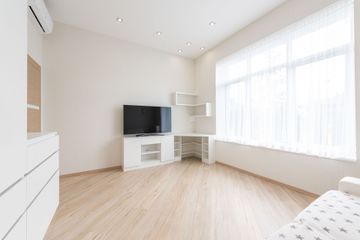 Bright minimalist living room with white walls, light wood floor, TV, and large window with sheer curtains