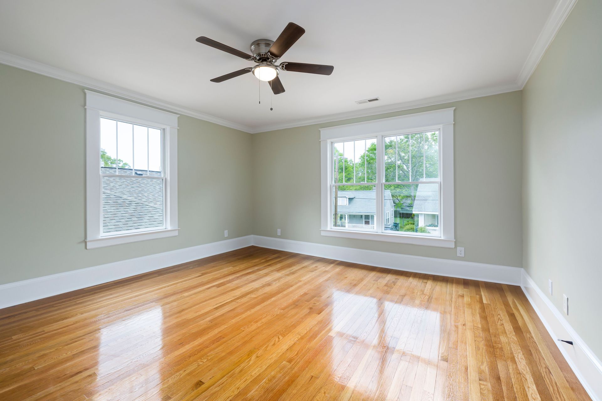 Empty living room with hardwood floors, light gray walls, ceiling fan, and large windows.