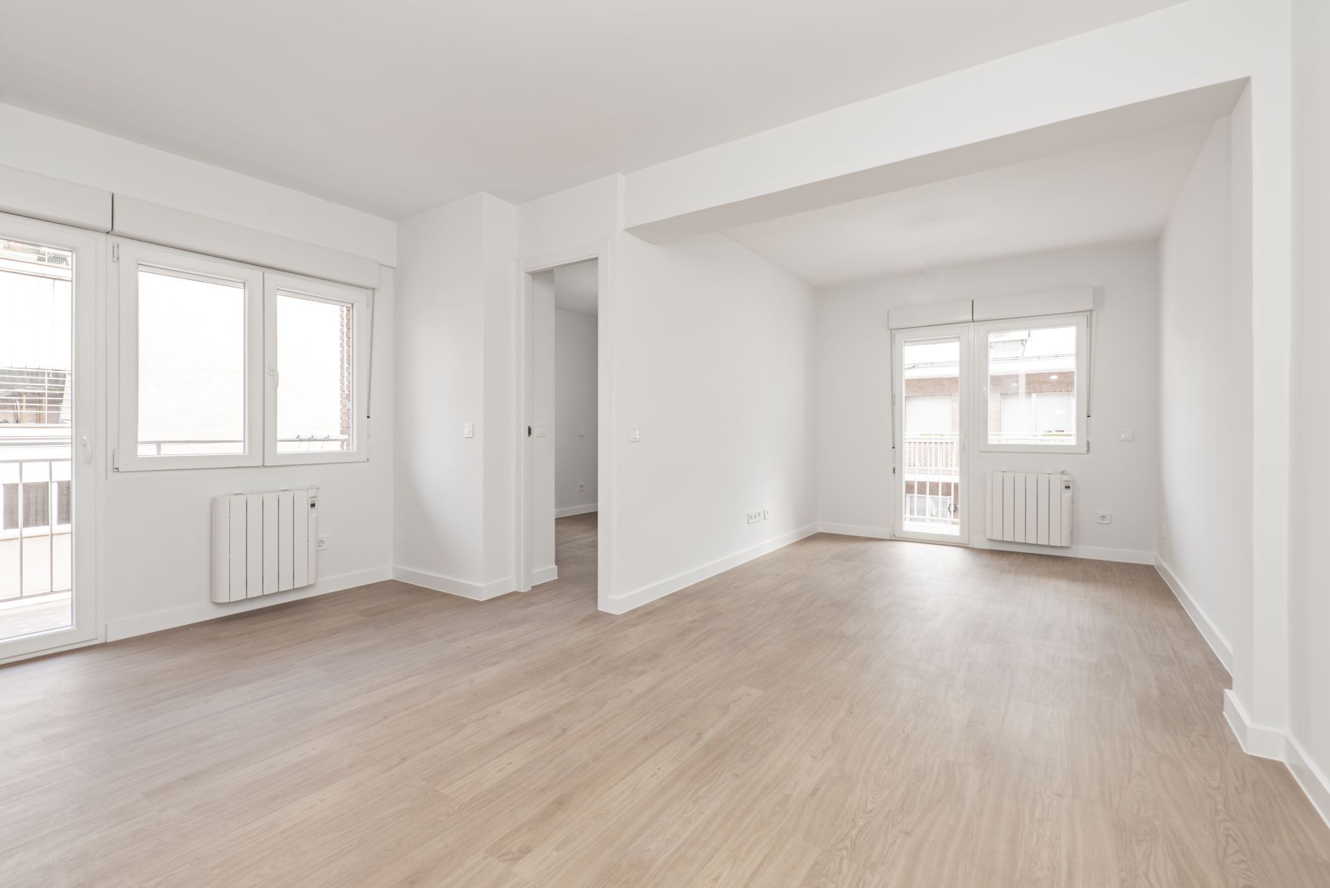 Bright empty apartment room with light wood floors, white walls, windows, and radiator.