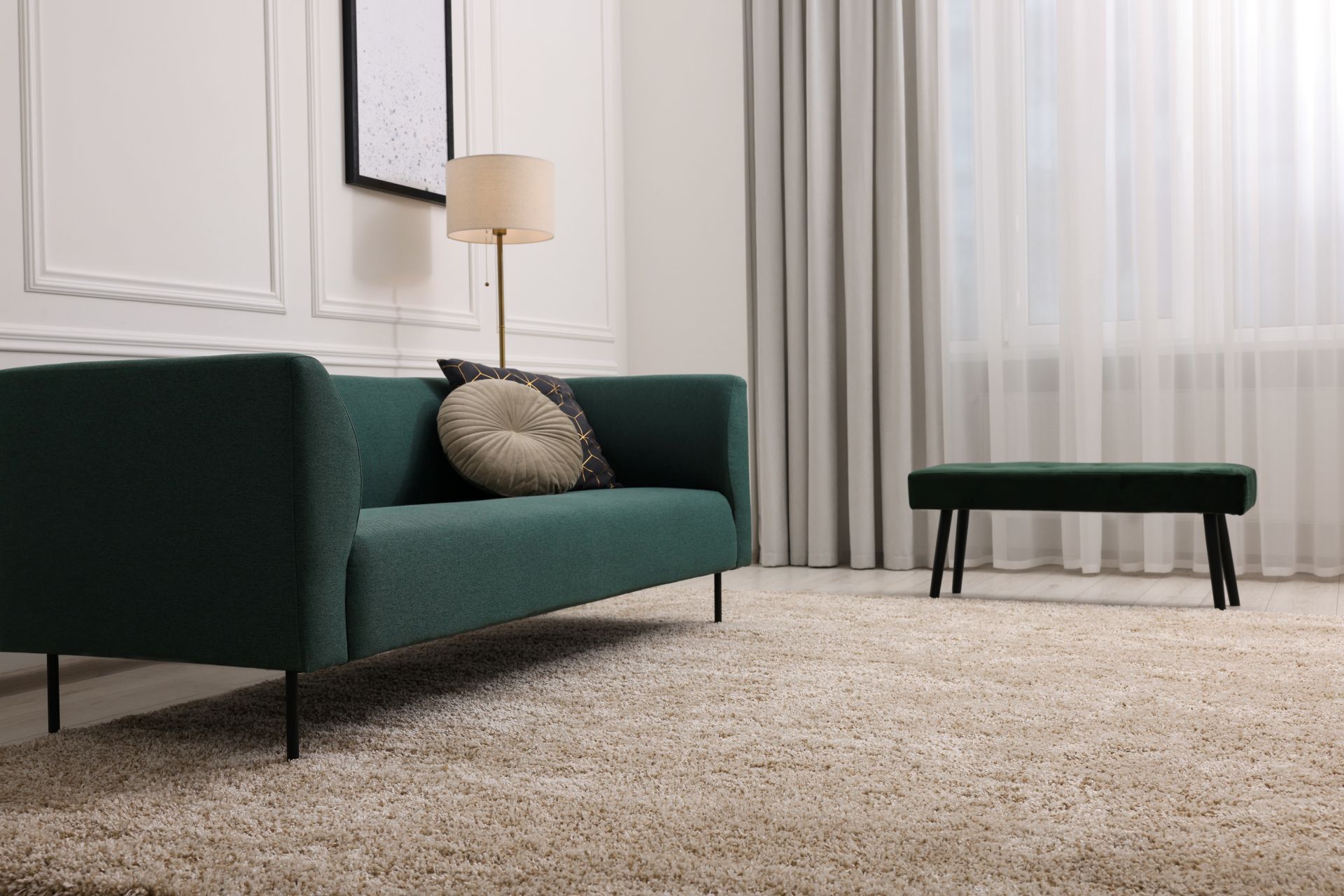Modern living room with a dark green sofa, lamp, bench, and sheer curtains on a light rug.