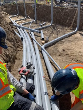 Two construction workers are working on a row of pipes in the dirt.