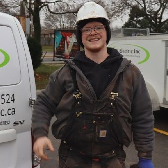 A man wearing a hard hat and overalls is standing in front of an electric inc van