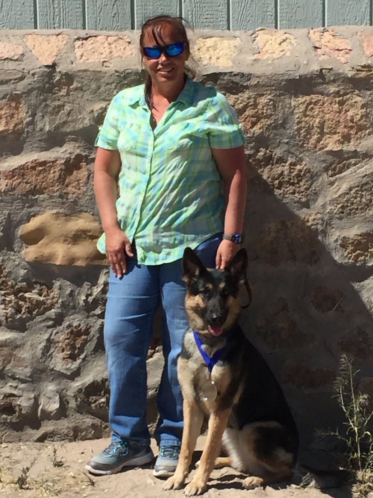 Woman in blue shirt and jeans with a German Shepherd dog; poses outside against a stone wall.