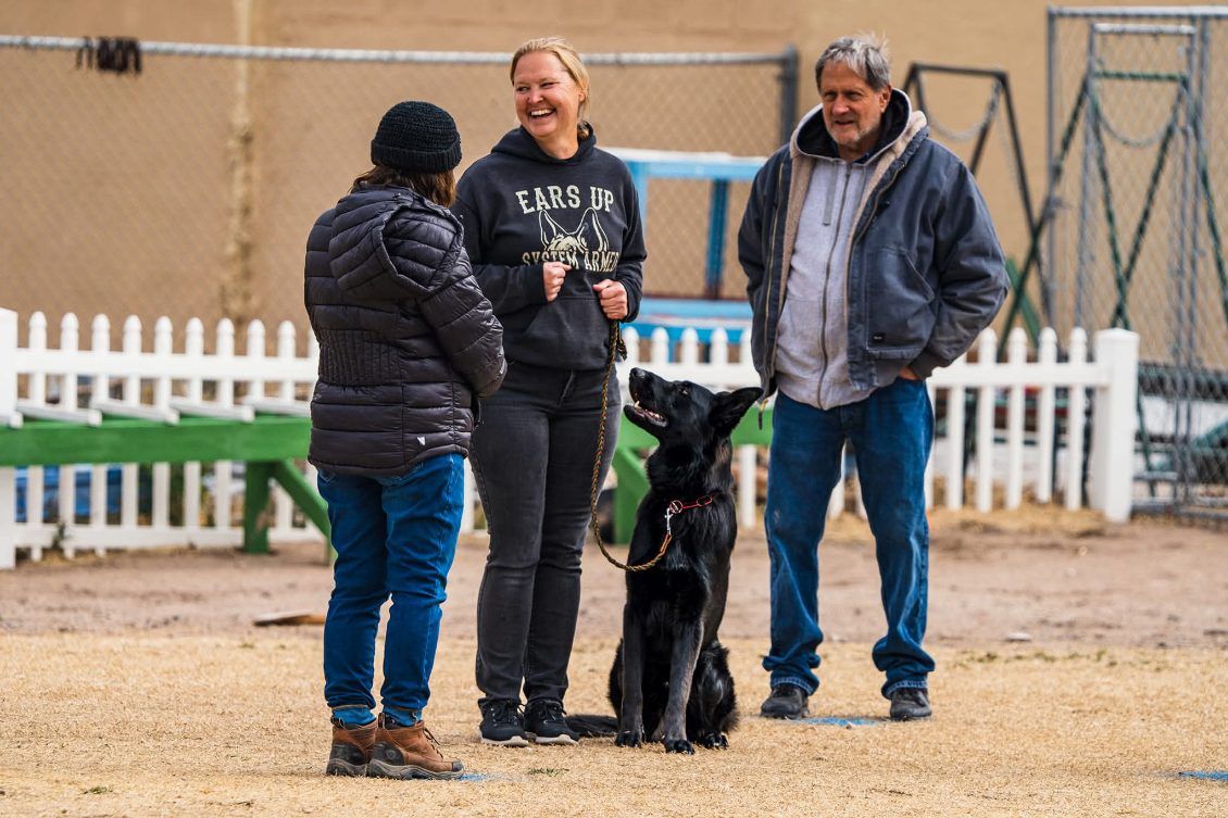 Three people with a black dog in a fenced outdoor training area, one person holding the dog’s leash.