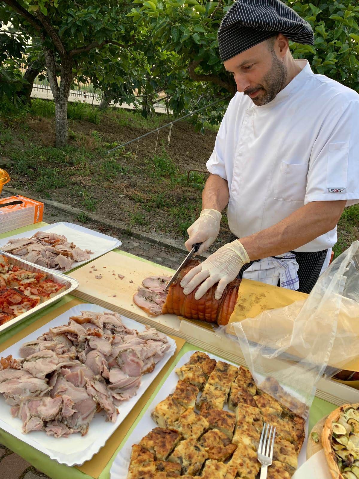 Un uomo affetta carne arrosto all'aperto accanto a vassoi di cibo preparato su un tavolo.