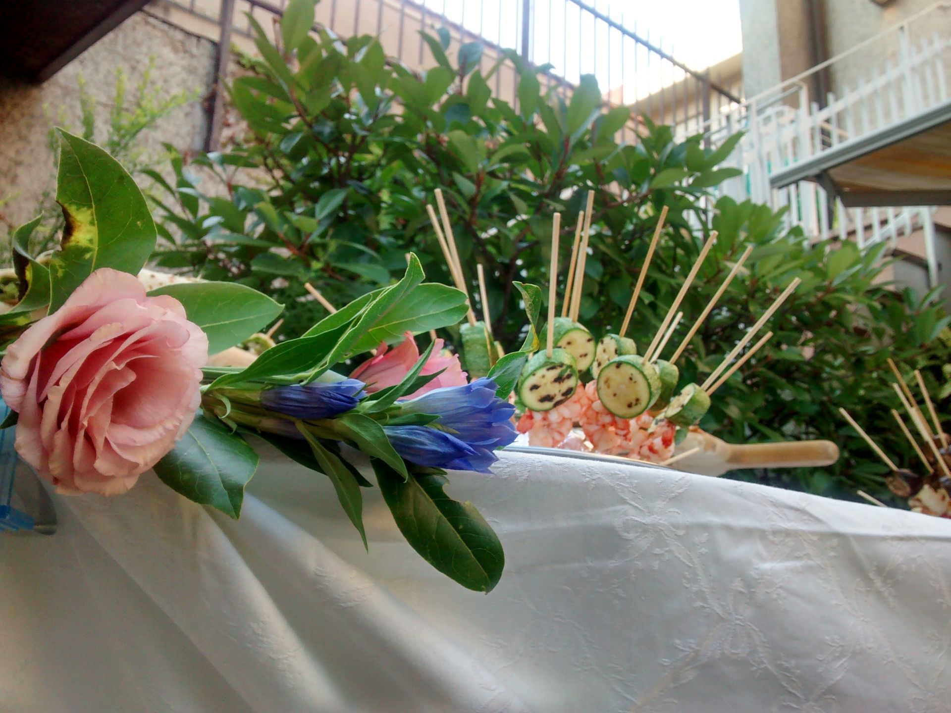 Composizione floreale con fiori rosa e blu su un telo bianco, posizionata all'aperto vicino a piante e scale.