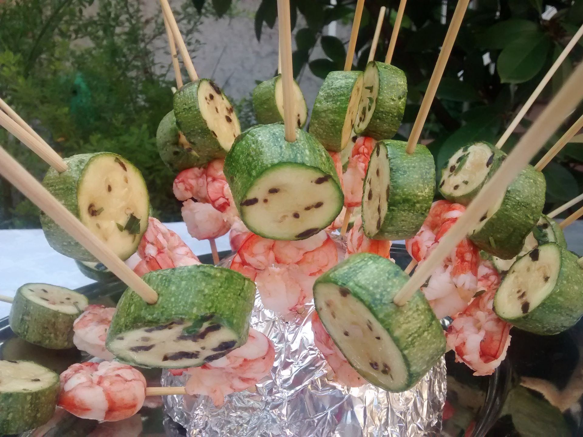 Antipasto di cetrioli e gamberetti infilzati su spiedini, disposto su un vassoio foderato di carta stagnola, con salsa all'arancia e guarnizione di foglie.