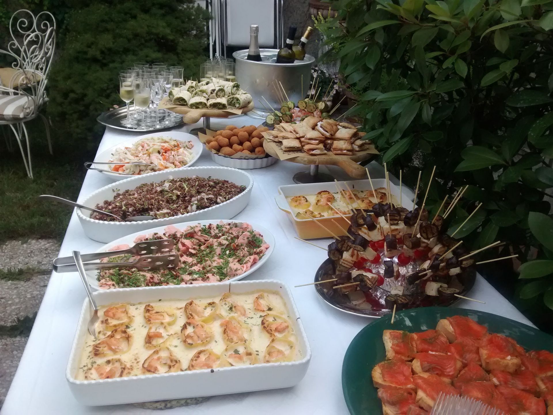 Tavolo da buffet all'aperto con un assortimento di antipasti, insalate, spiedini e dessert, apparecchiato su una tovaglia bianca.
