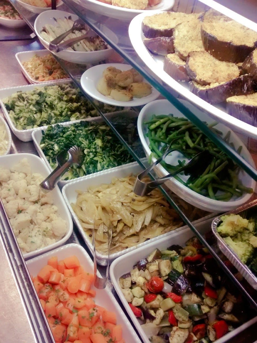 Buffet di insalate con un assortimento di piatti pronti in vassoi bianchi, tra cui verdure a foglia verde, ortaggi, pasta e fritti.
