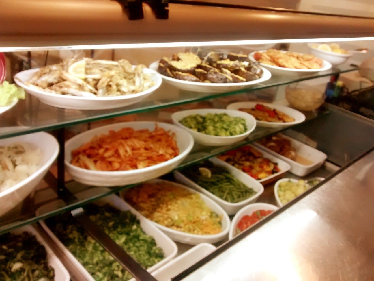 Bancone buffet con insalate assortite e contorni in ciotole bianche, illuminato da una luce calda.