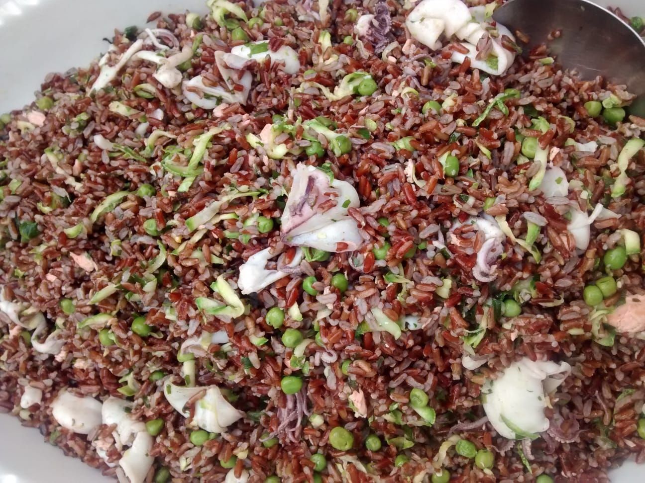 Primo piano di un'insalata mista di cereali con riso rosso, piselli, erbe aromatiche tritate e pezzi di pesce o frutti di mare.