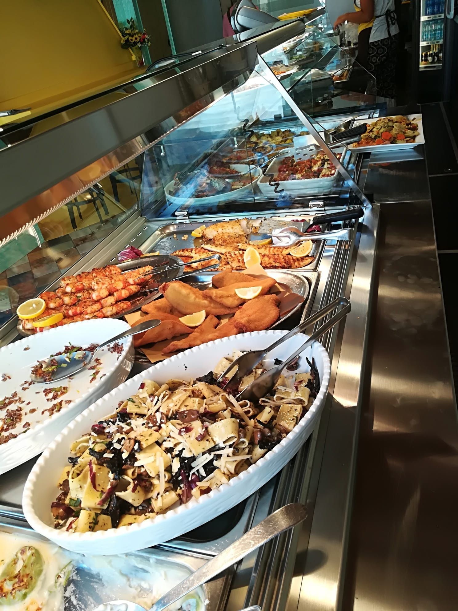 Bancone buffet con assortimento di piatti caldi e insalate sotto vetrina, vassoi da portata in acciaio inox.