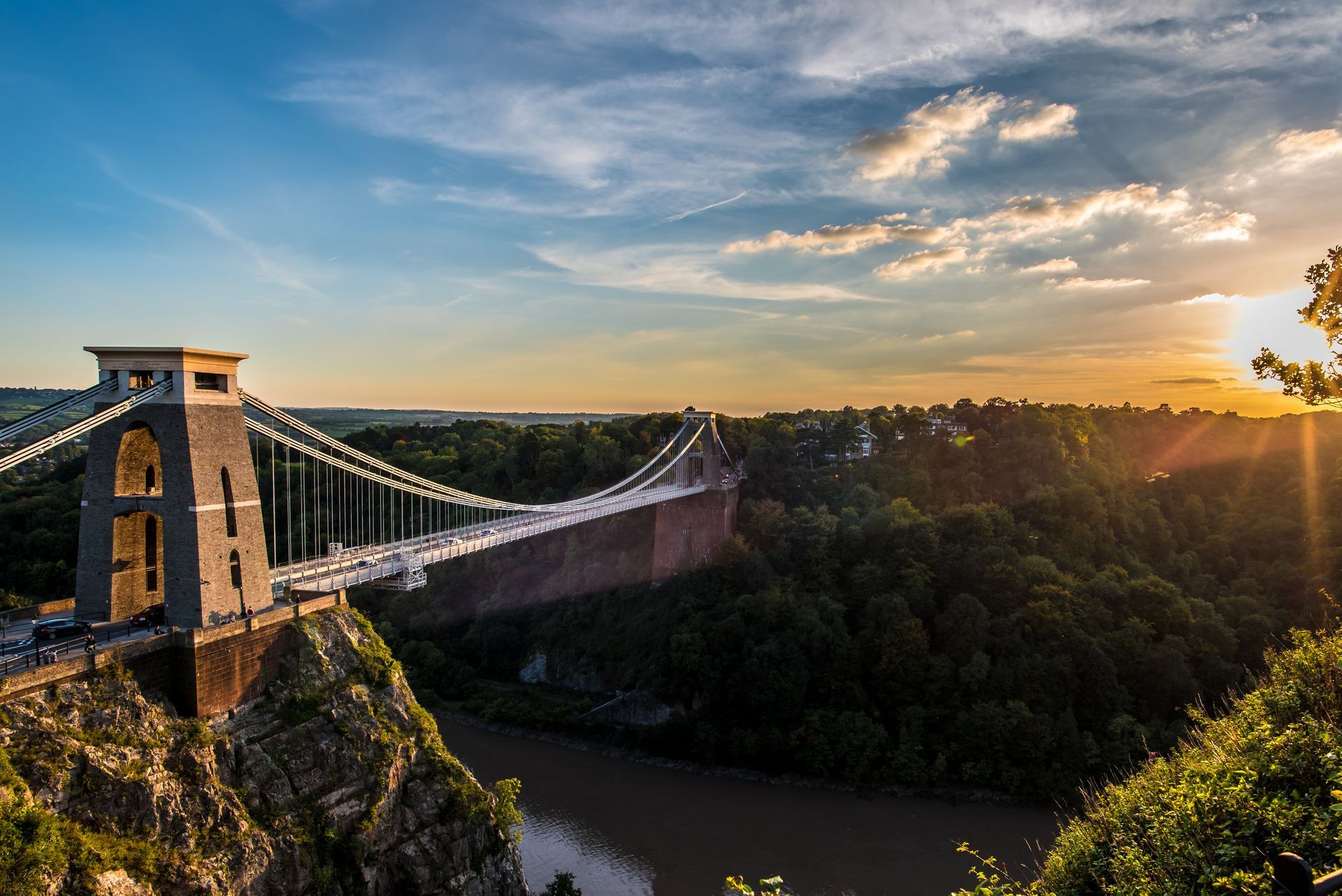 A bridge over a river at sunset with the sun shining through the clouds.