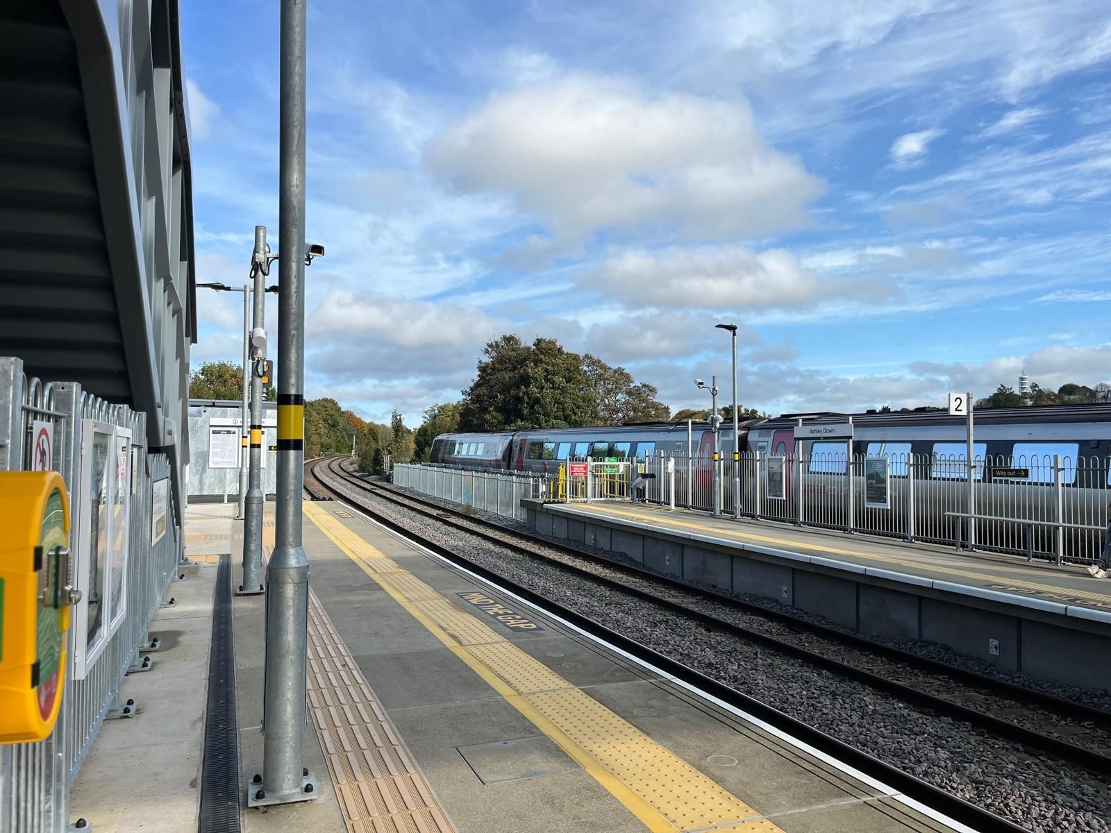 Train station platform with tracks, overhead structure, and trains in the distance under a blue sky.
