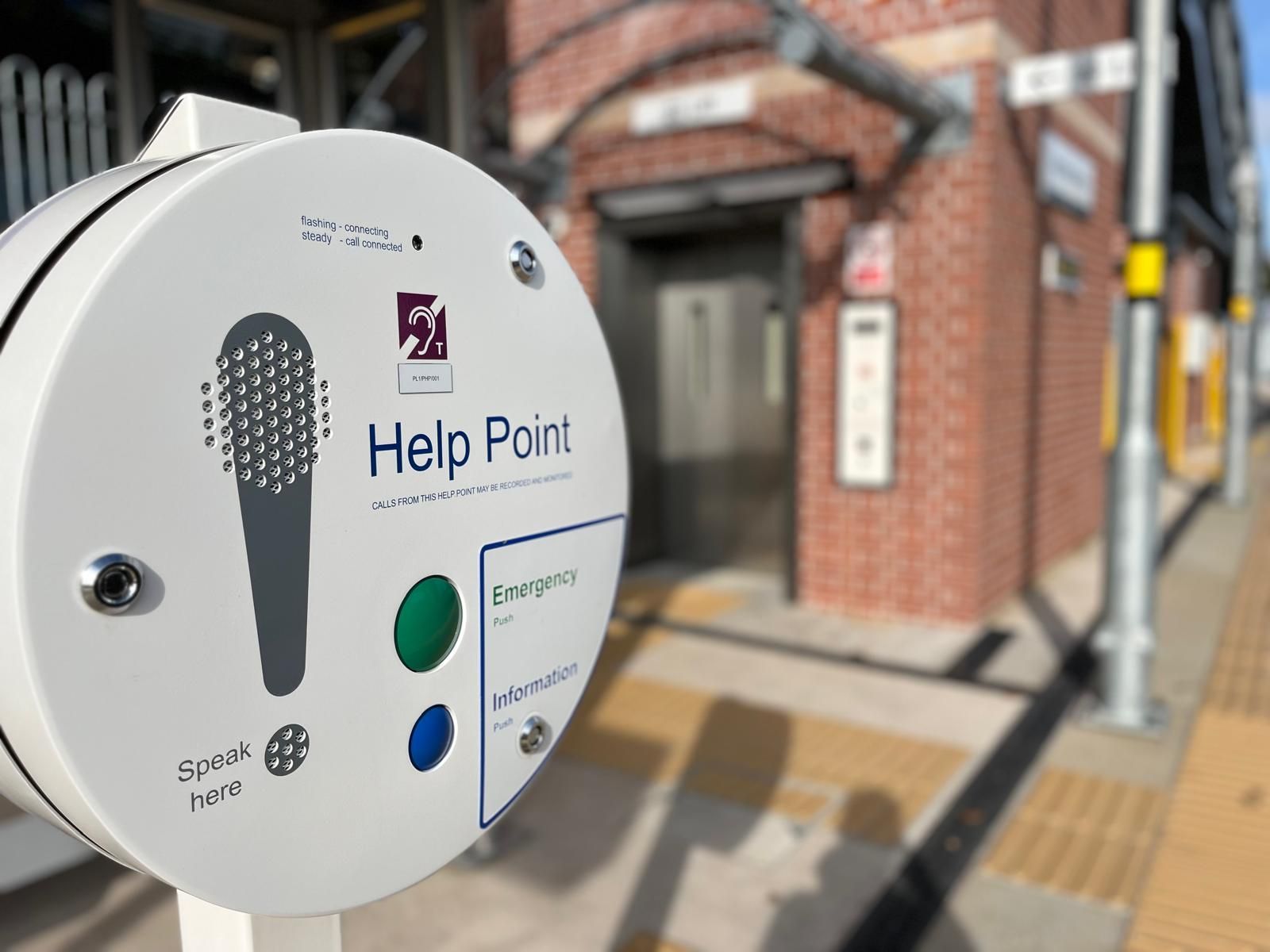 Help point station with microphone symbol, emergency and information buttons. Brick building in background.