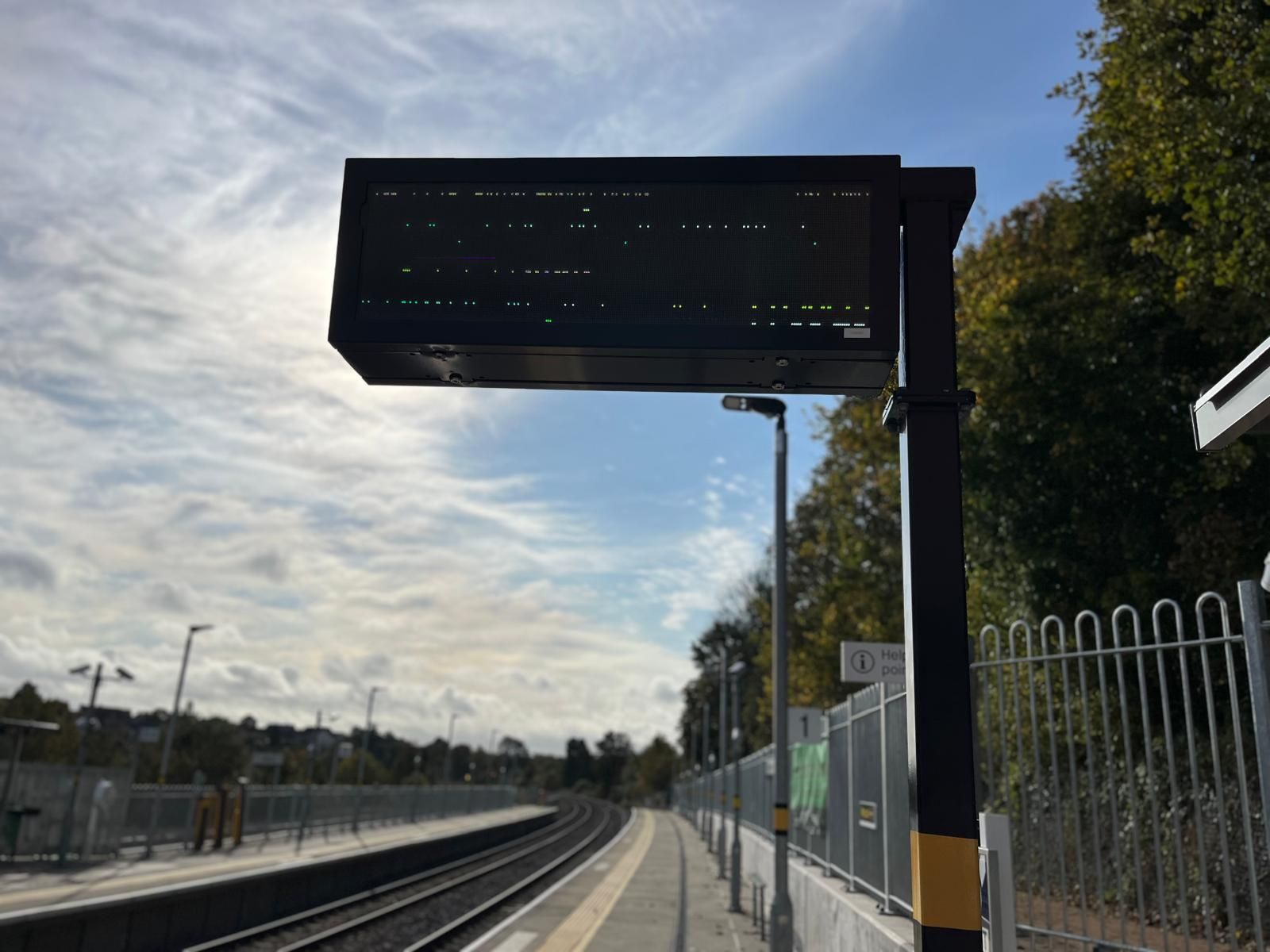 Train station departure board atop a black pole; tracks, platform, and sky in background.