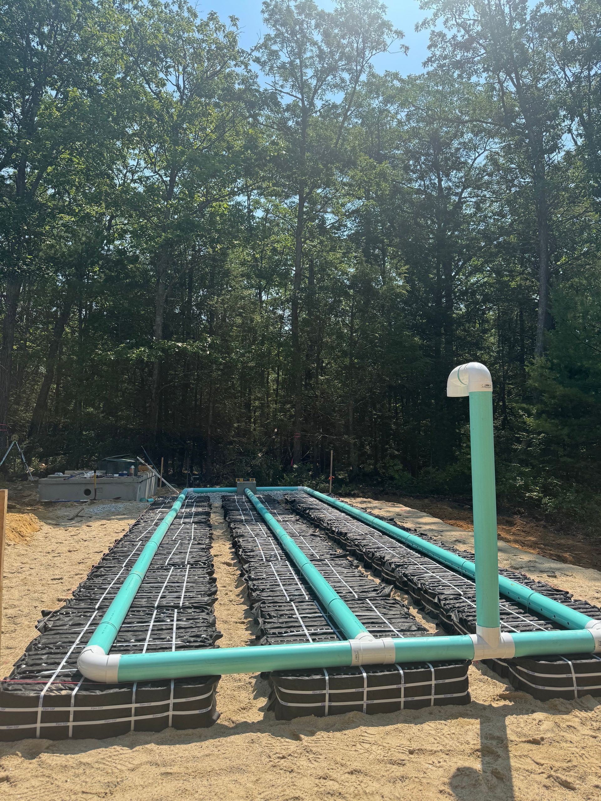 PVC pipes laid in a gravel area beside a wooded tree line, with a white vent pipe in front