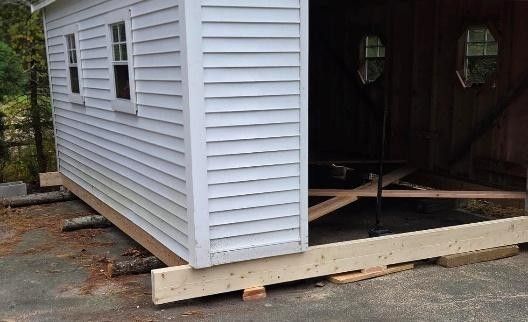 White shed or small building on wood skids, partially installed on a driveway with open doorway.