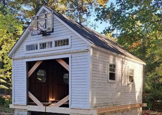 Small white wooden shed with open front and gabled roof in a wooded yard