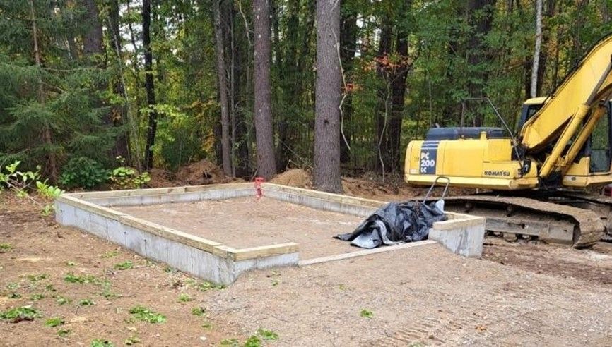 Concrete slab foundation under construction beside a yellow excavator in a wooded area.
