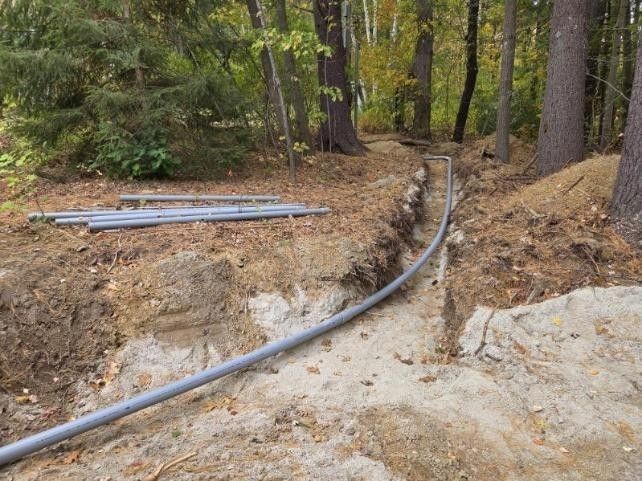 Gray hose running through a muddy wooded trench beside stacked metal pipes and trees