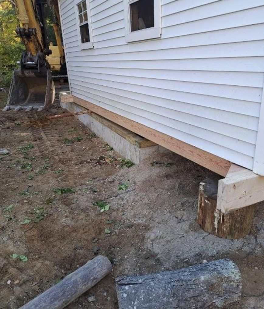 House exterior with raised siding, exposed foundation, and an excavator beside a dirt yard.