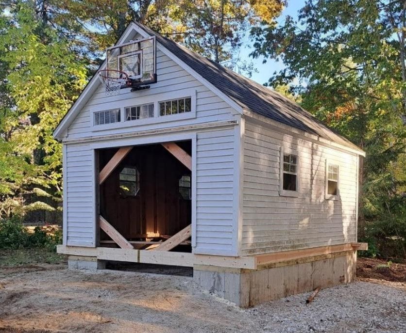 Small white shed with open front, set on a gravel site among trees in daylight.