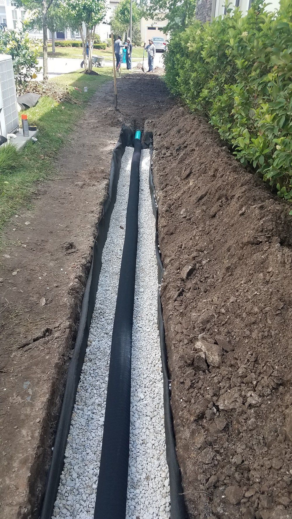 A narrow gravel trench under construction beside a hedge, with soil piled on both sides.