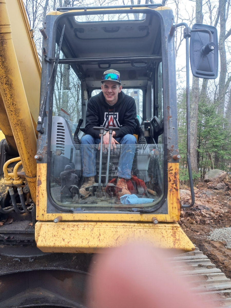 Person smiling inside a yellow excavator cab on a muddy work site.