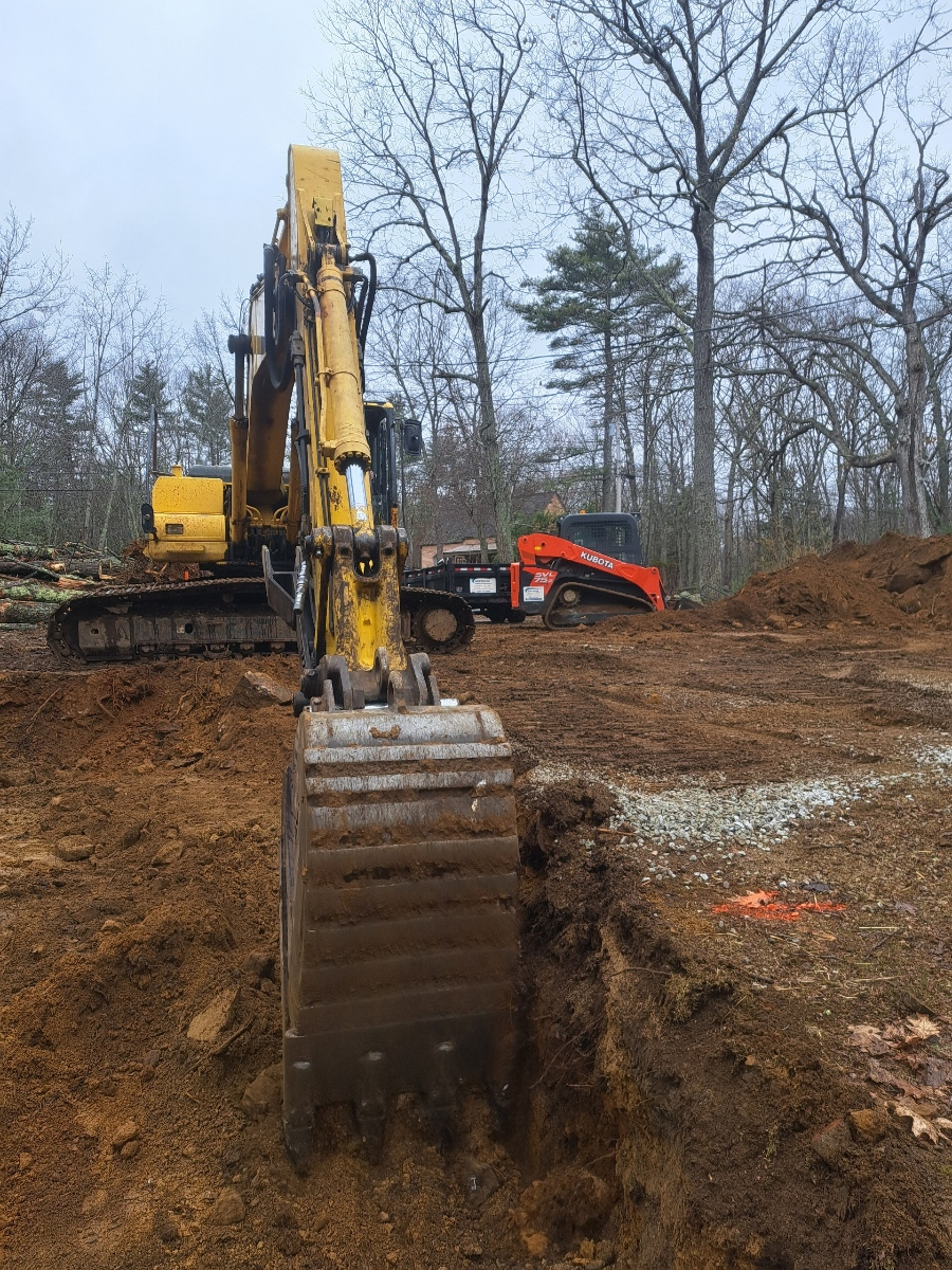 Yellow excavator with raised bucket on a muddy construction site, with bare trees and an orange mini excavator in back.